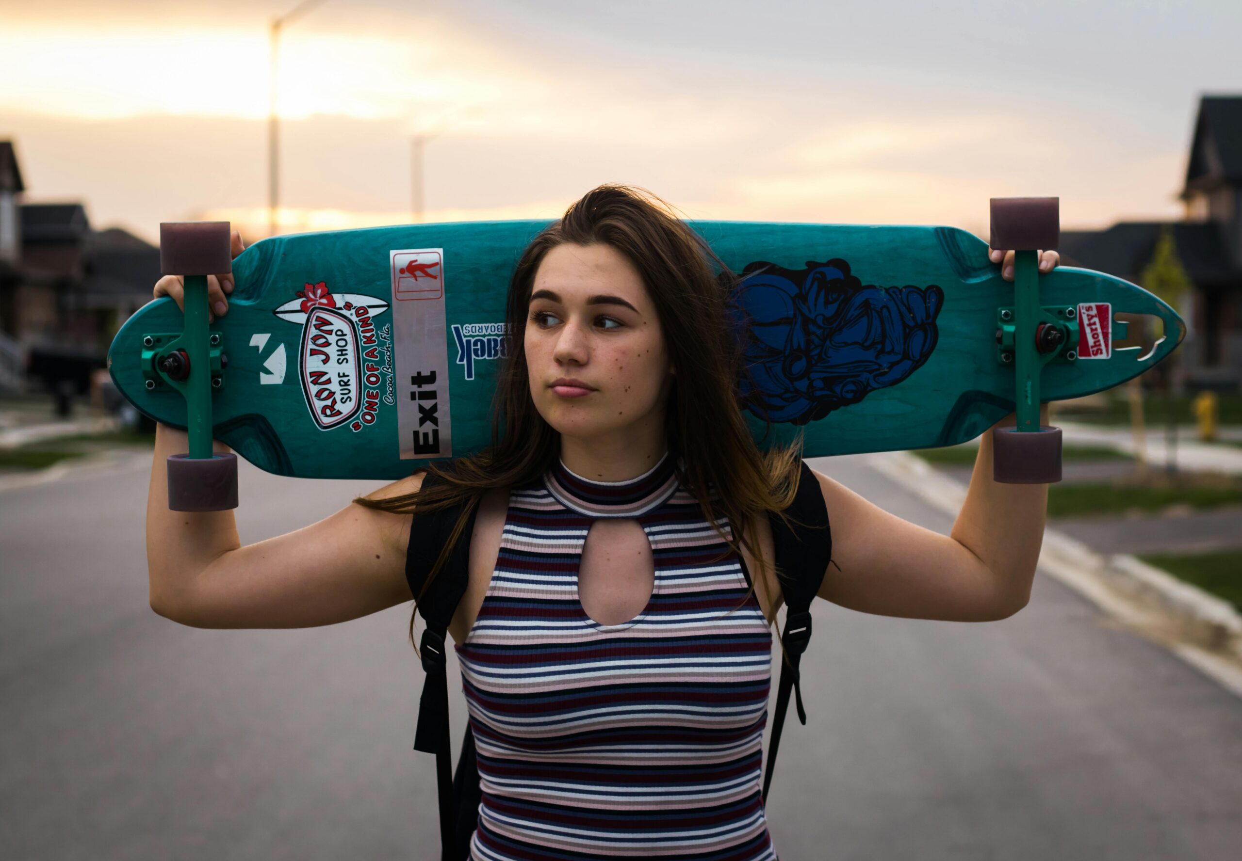 Teen girl carries a longboard skateboard on shoulders in suburban street at sunset, showcasing modern skate style.